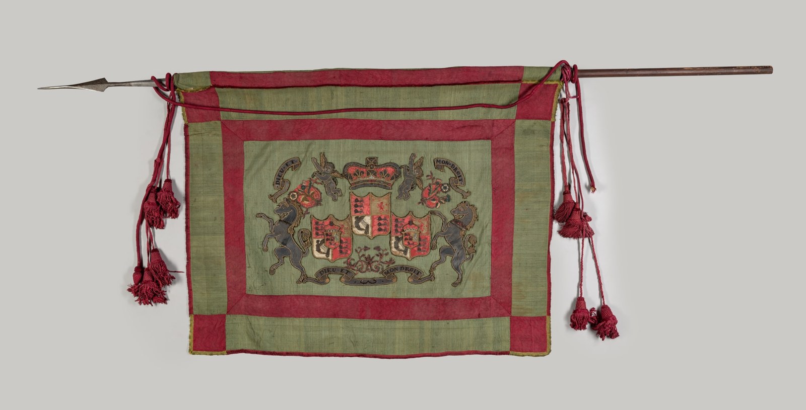 A green rectangular textile with a broad red border hangs from a pole. An embroidered achievement occupies the centre, with flanking figures and scrolling elements around the shield.