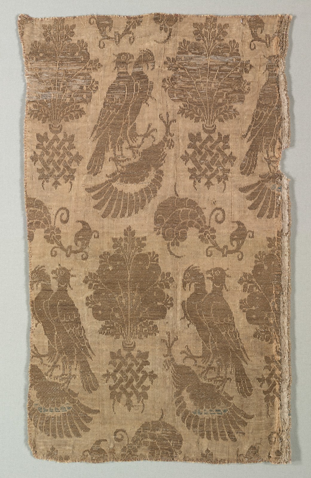 Large paired bird forms alternate with upright plumes and curling ornaments in a gold-patterned fabric. The design repeats across the surviving textile.