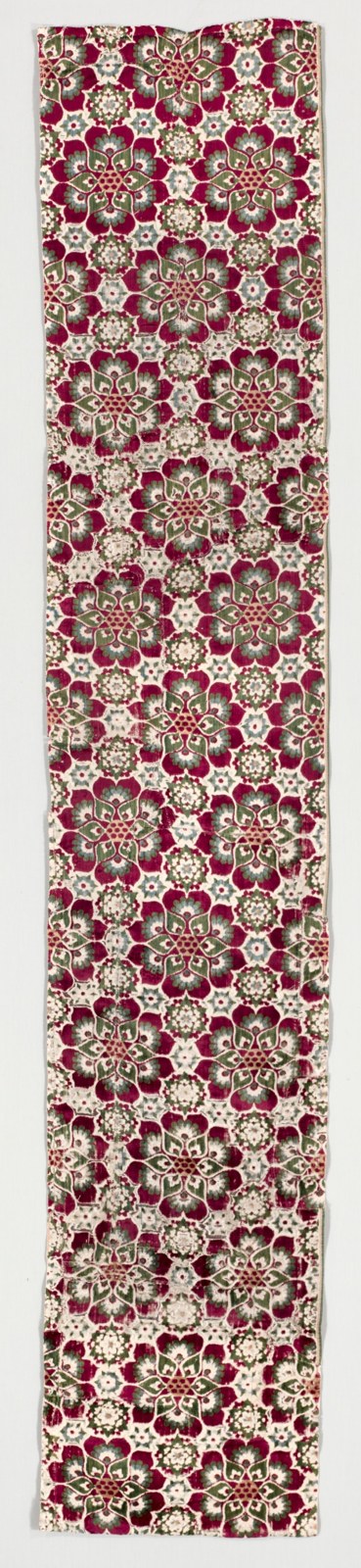 A long, narrow textile fragment repeats large red-and-green rosettes against a pale ground. Small groups of round forms sit inside the repeated centres.