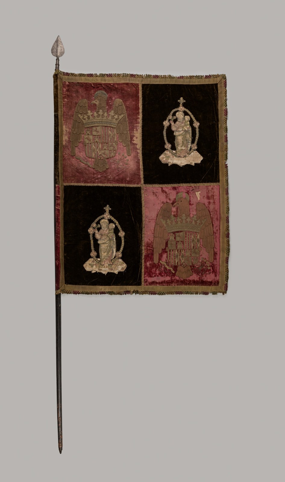 Four large textile compartments alternate two armorial displays with two images of a mother and child. Fringe outlines the rectangular fabric, which hangs from a long pole.