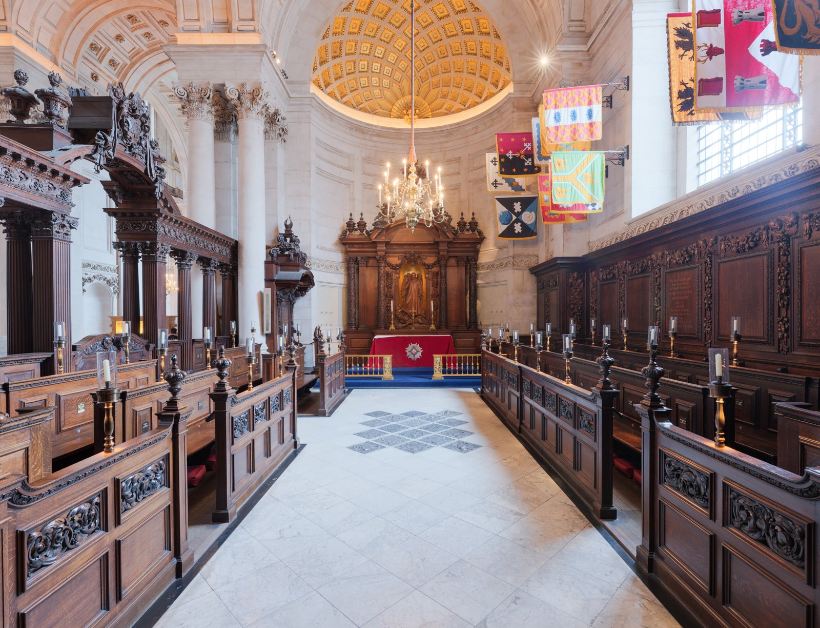 Coloured rectangular banners hang over the stalls on the right of a bright chapel. Carved wooden seats and wall panels face one another across an aisle leading to an altar.
