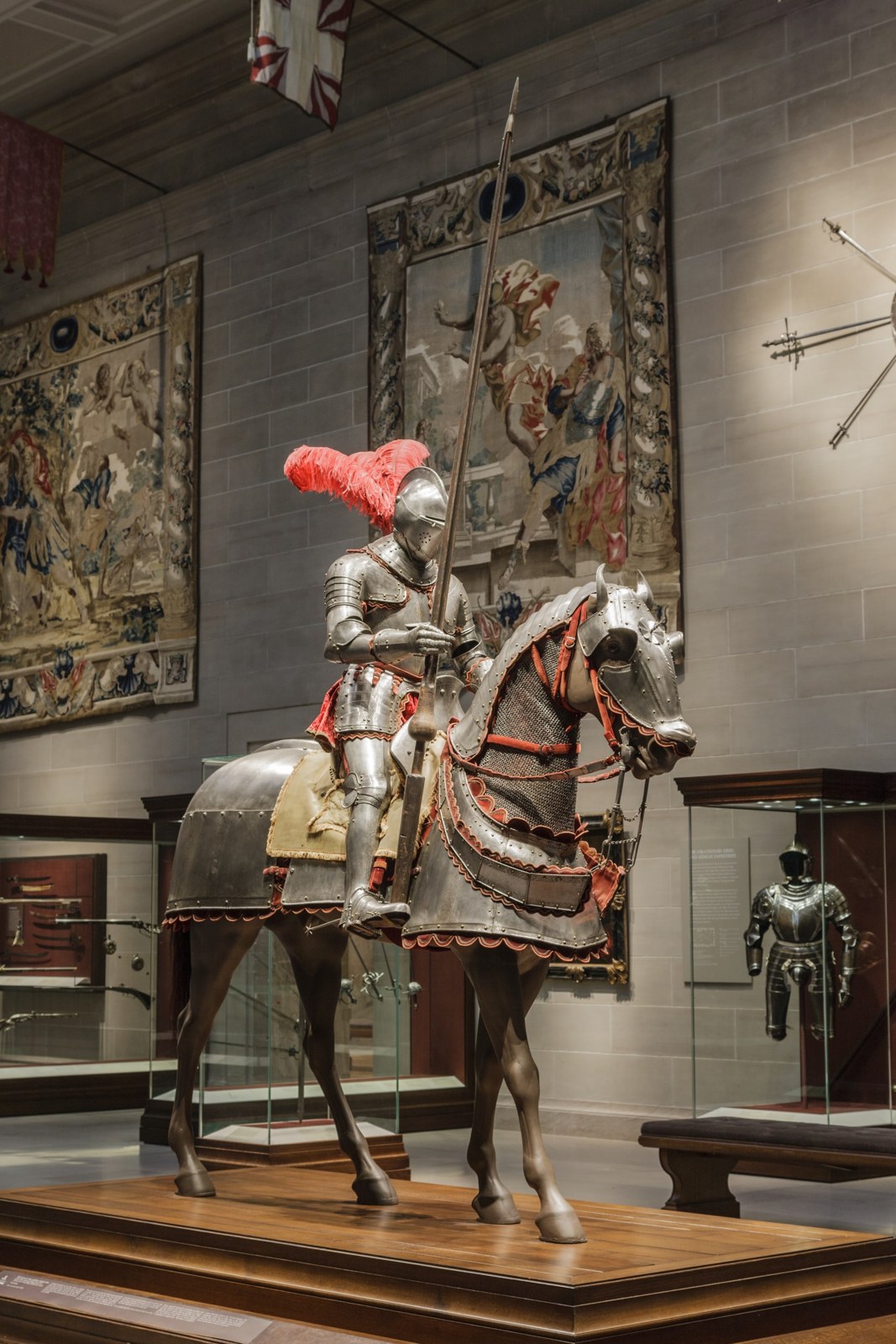 A fully armoured rider sits above an armoured horse. Red fabric, etched metal, and the museum’s supporting mount make this a much larger display than a single painted shield.